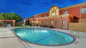 A swimming pool with a tiled deck, surrounded by a building with arched windows and a red facade.