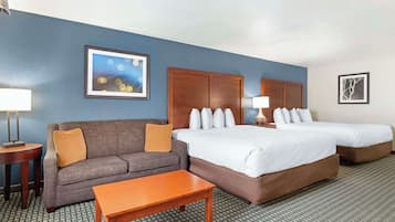 Habitación de hotel con un sofá gris, una mesa de madera, dos camas con sábanas blancas, una alfombra estampada y dos cuadros en las paredes.