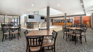 A spacious dining area with multiple tables and chairs, a kitchen area in the background, and a high ceiling with recessed lighting.