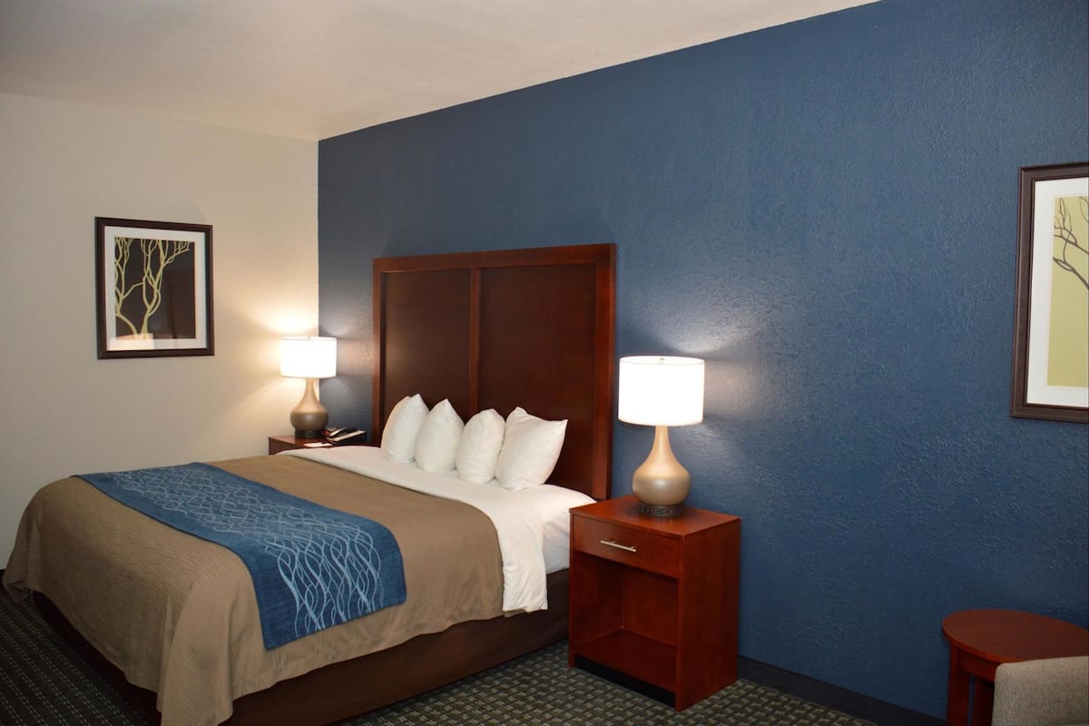 A hotel room with a bed, two bedside lamps, a nightstand, a small table, and two framed pictures on the walls.