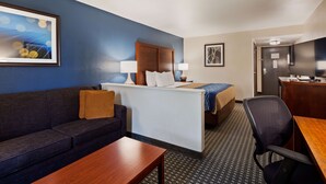 A hotel room with a dark blue wall, a wooden headboard, a bed with a blue and beige blanket, a white bedside table, a wooden desk, a black office chair, and a framed picture on the wall.
