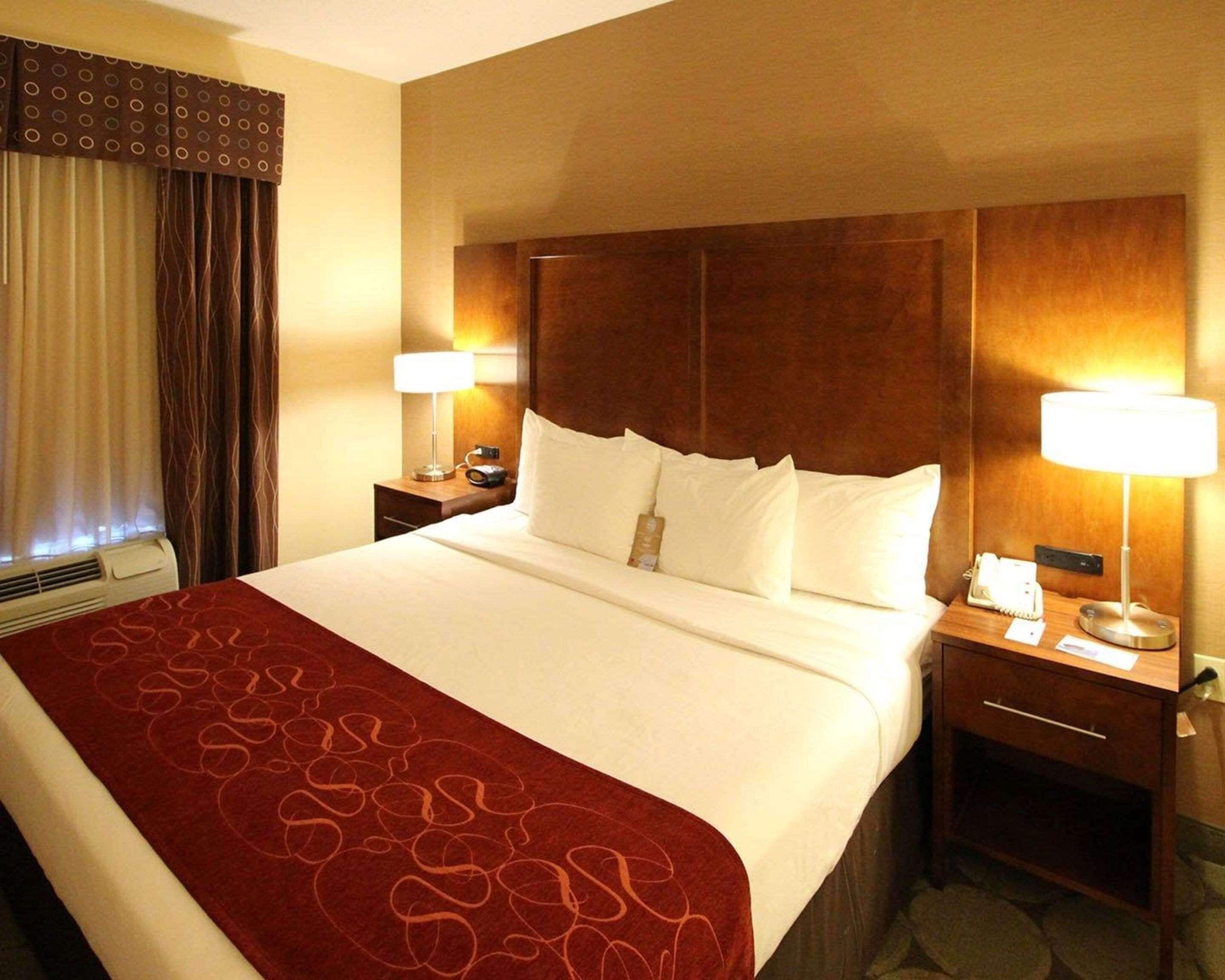 A hotel room with a large bed, two bedside tables, a wooden headboard, a red patterned bedspread, and two lamps.