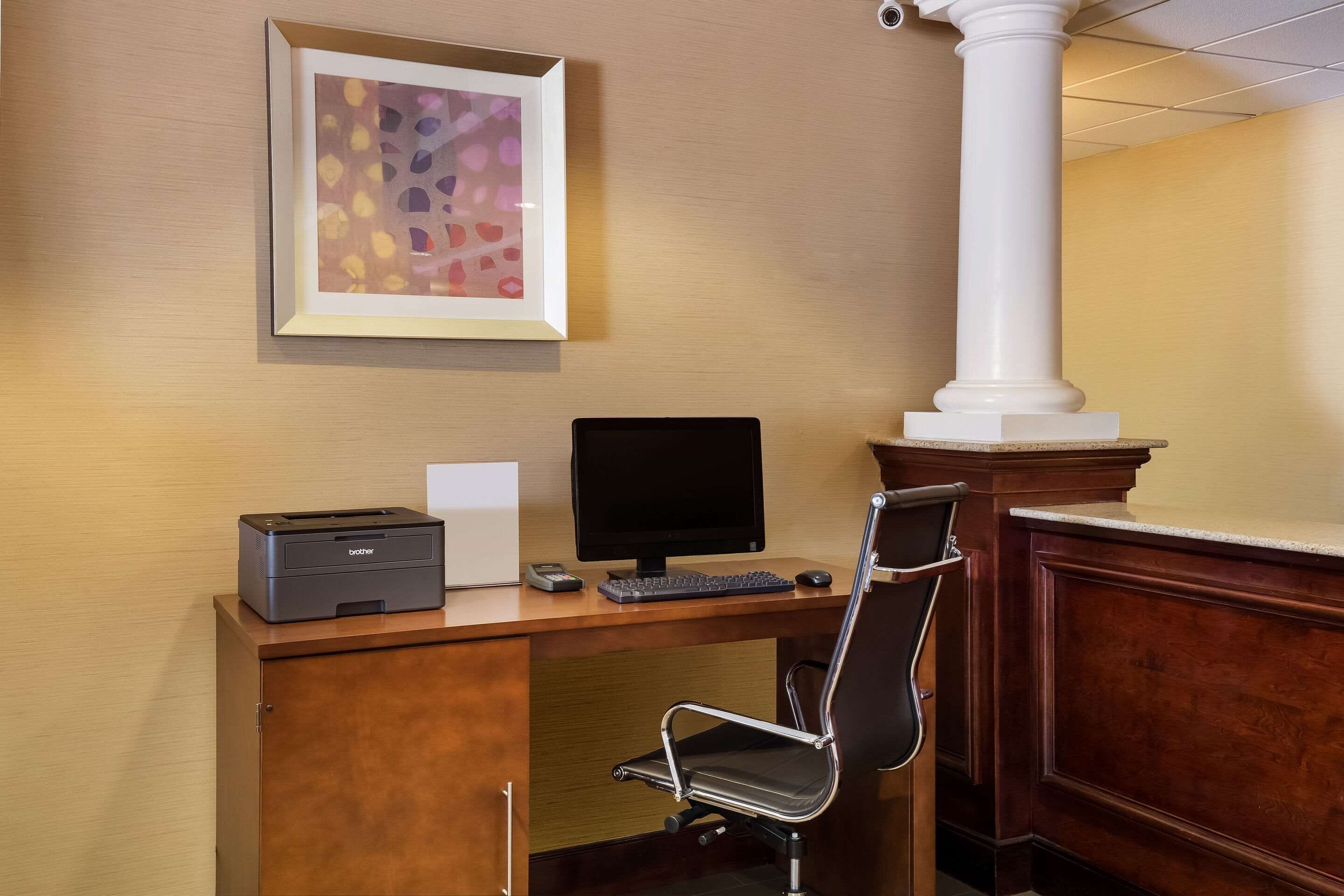 A wooden desk with a printer, monitor, and keyboard, a chair, and a framed abstract painting on the wall.