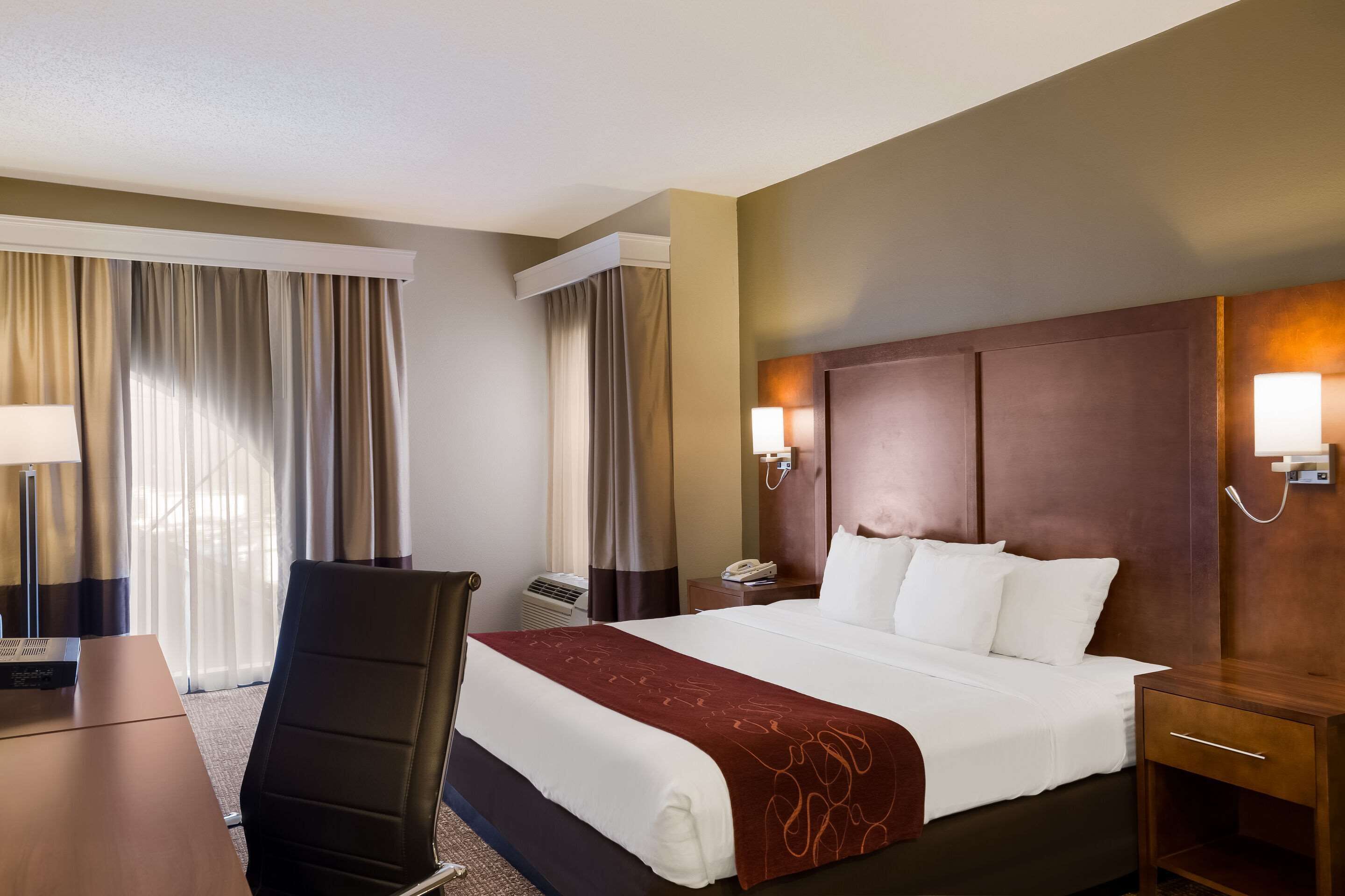 A hotel room with a large bed, a desk, a chair, a nightstand, and a lamp.