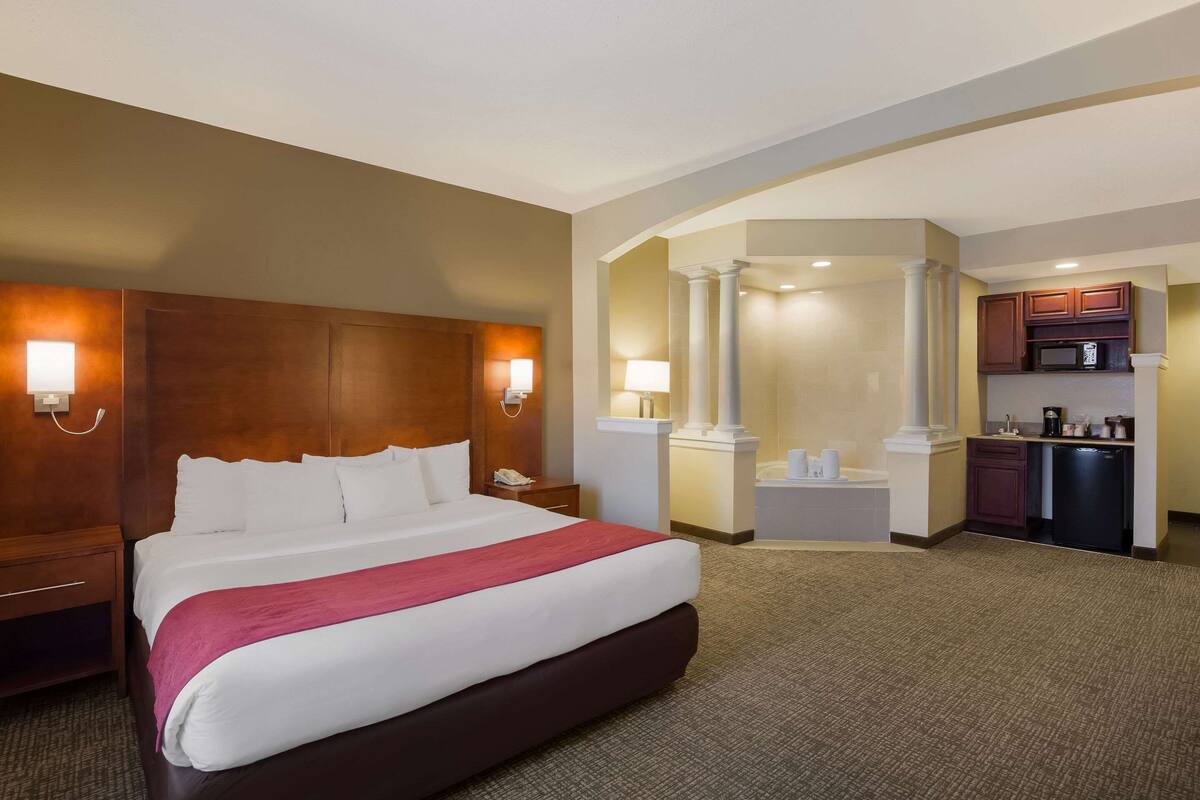 A hotel room with a large bed, bedside lamps, a small desk, and a kitchenette area.