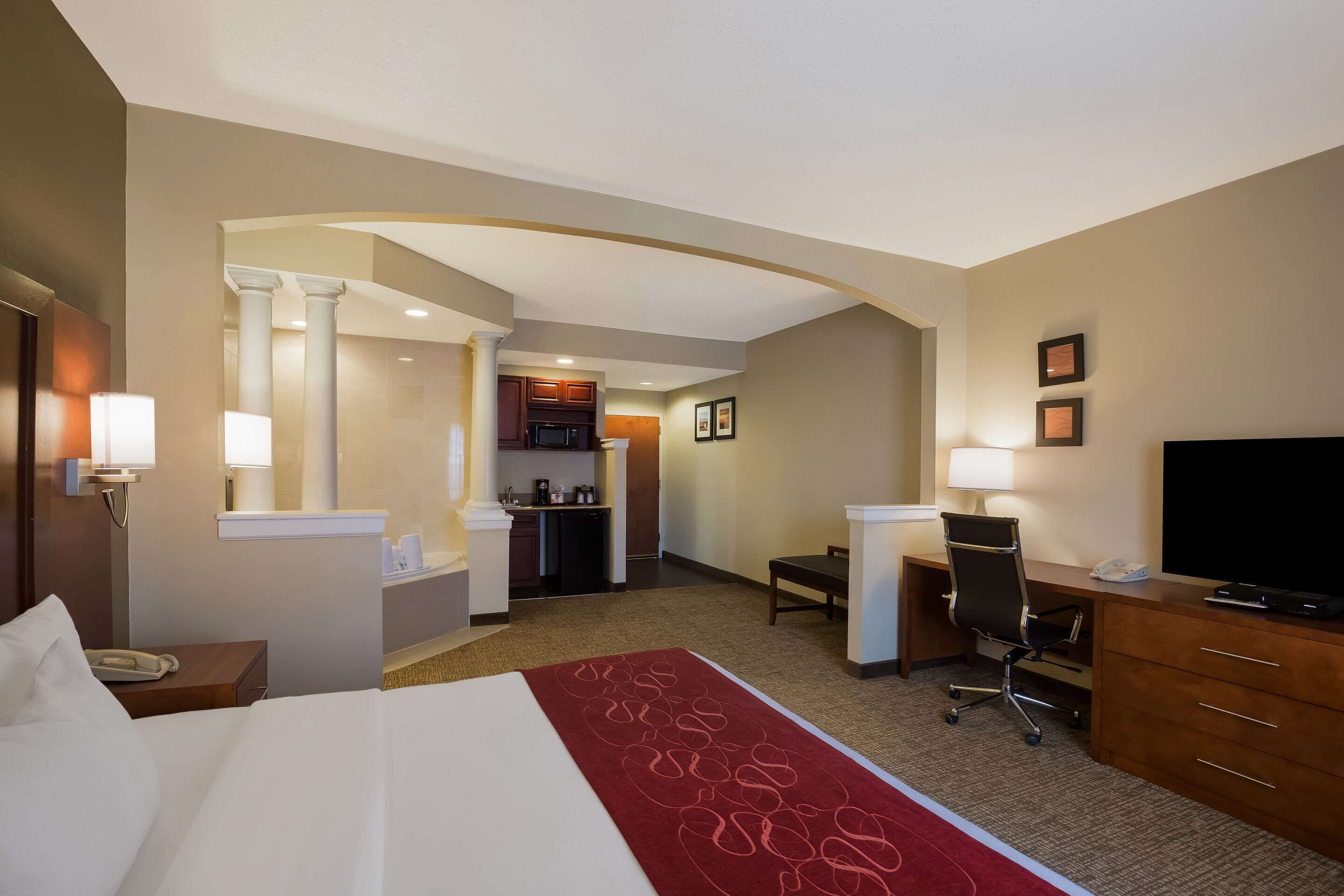 Habitación de hotel con cama, escritorio, silla, televisión y una kitchenette al fondo.