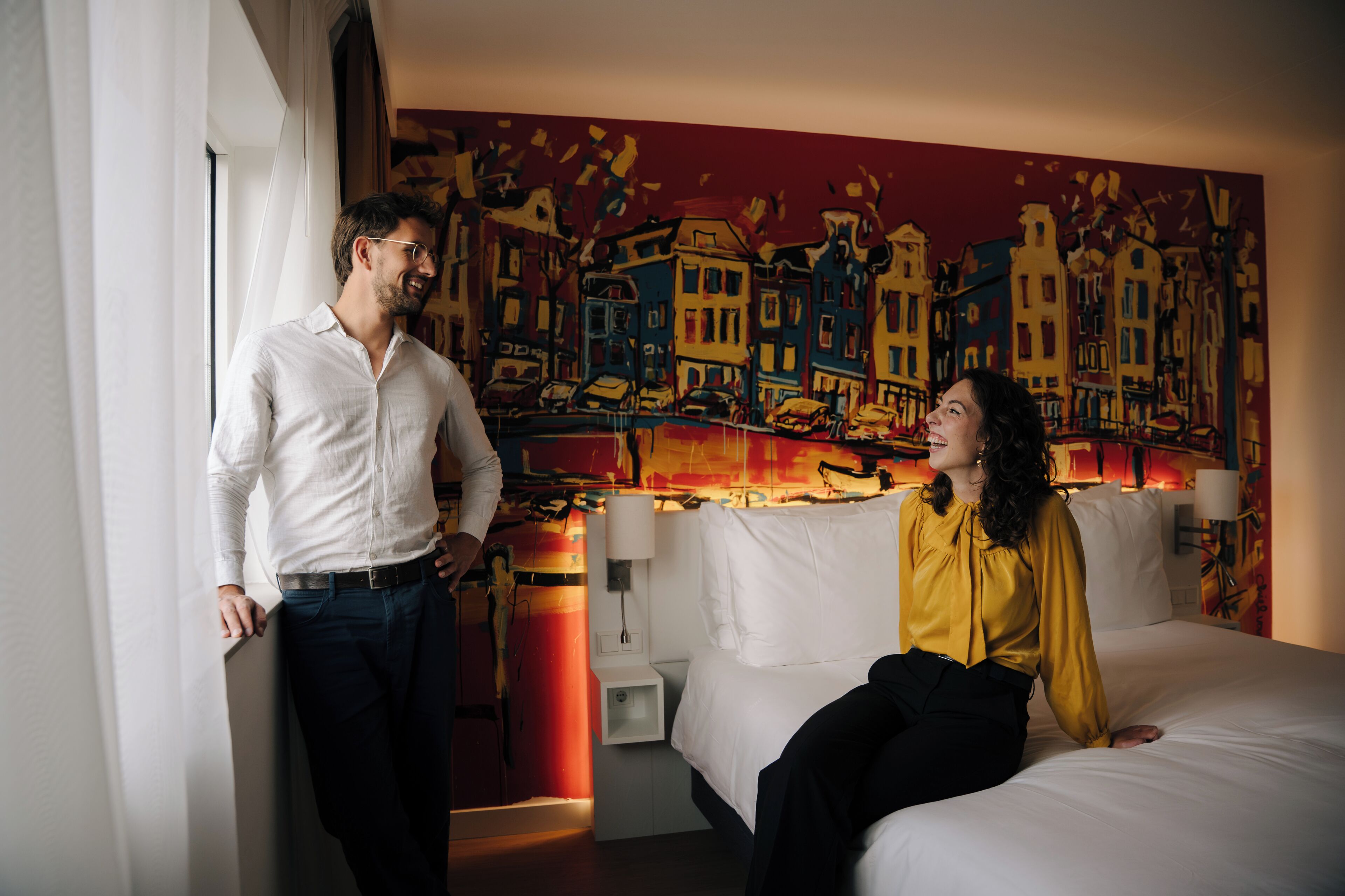 A hotel room with a bed, a painting of a cityscape, and a nightstand with a lamp.