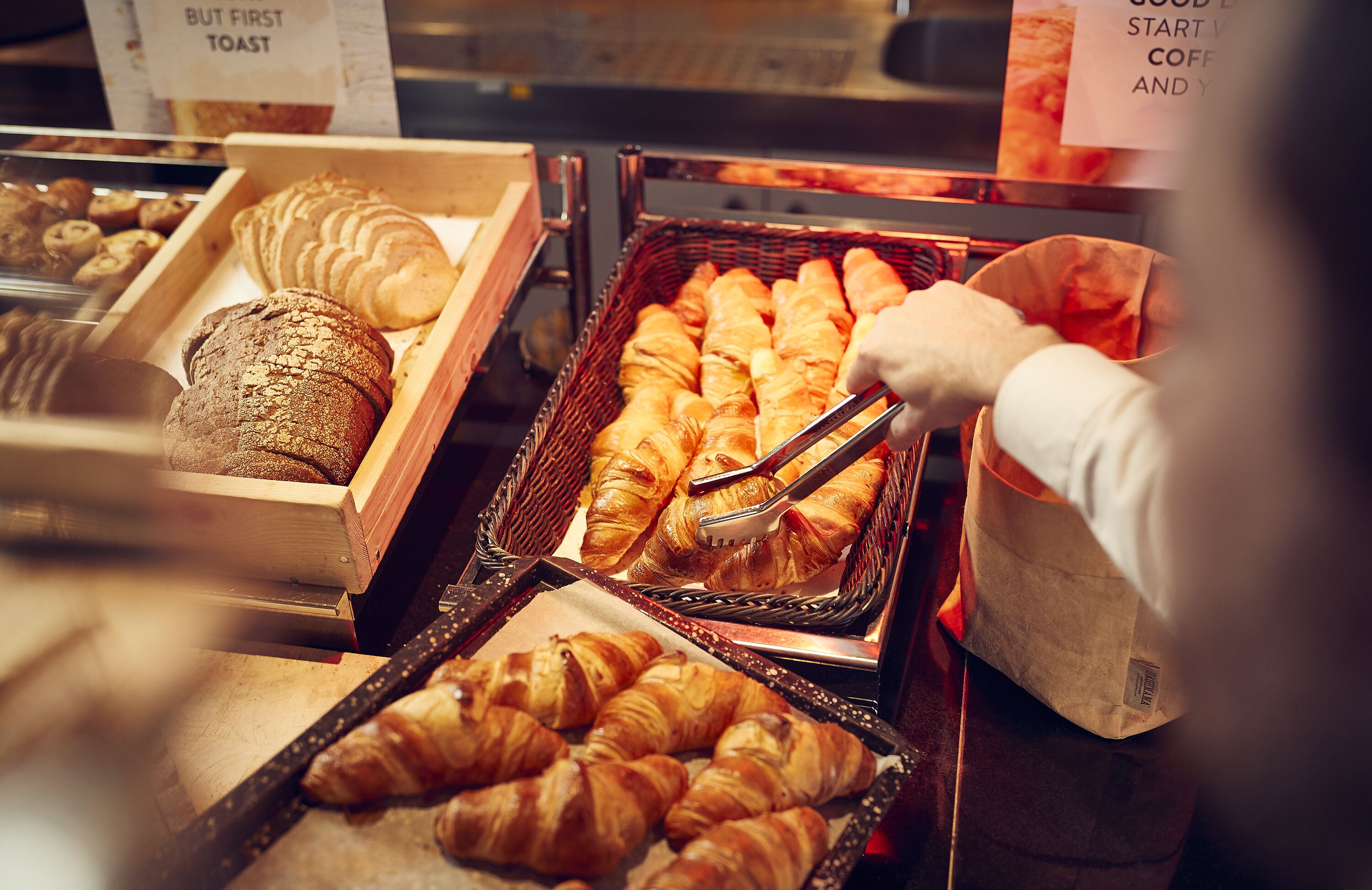 Leipomotuotteiden esillepano, jossa on erilaisia leivonnaisia, kuten croissantteja ja leipää, ja henkilö, joka käyttää pihdeillä tuotteiden käsittelyyn.