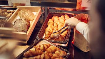 Leipomotuotteiden esillepano, jossa on erilaisia leivonnaisia, kuten croissantteja ja leipää, ja henkilö, joka käyttää pihdeillä tuotteiden käsittelyyn.