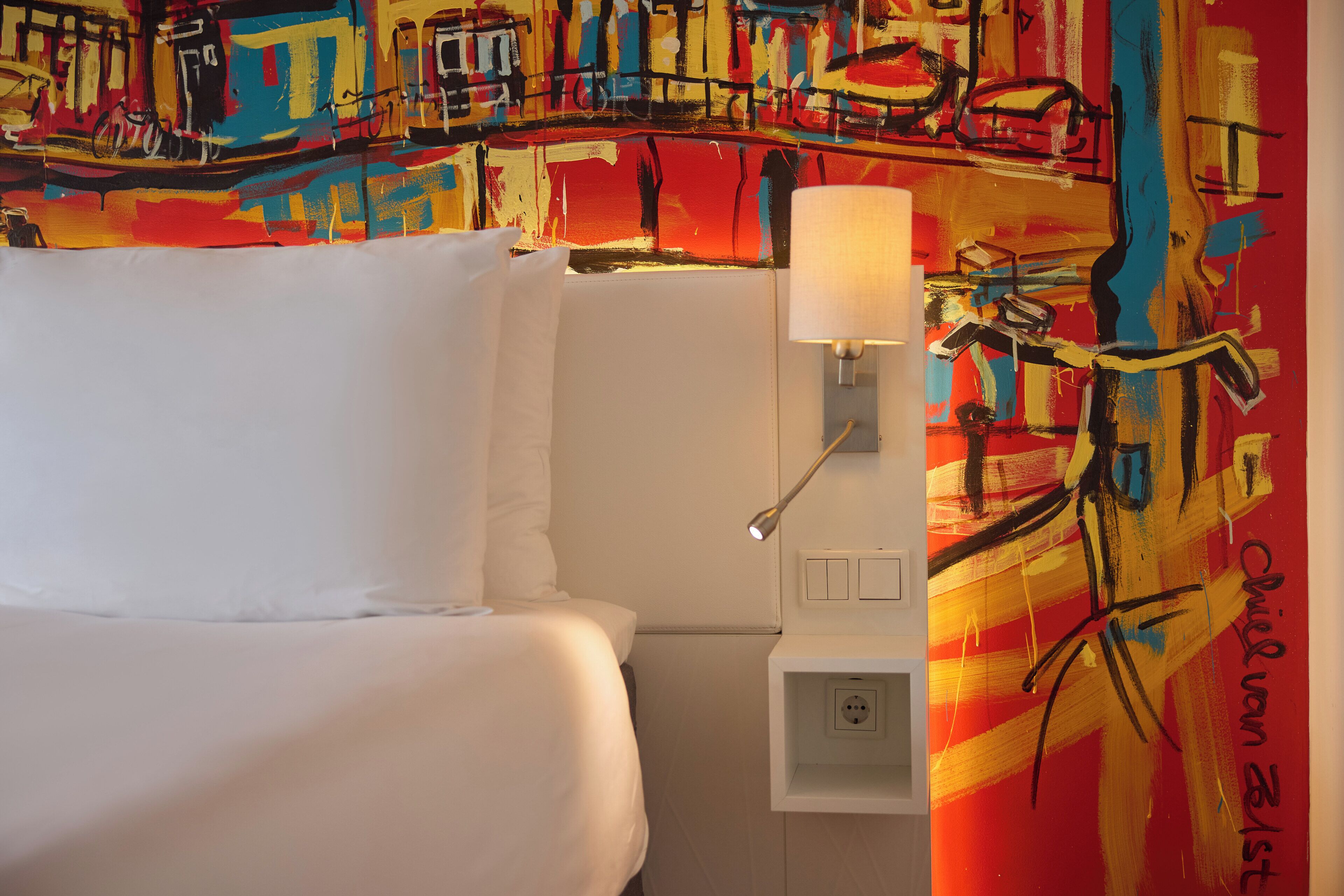 A hotel room with a bed, a nightstand, a lamp, and a colorful abstract painting on the wall.