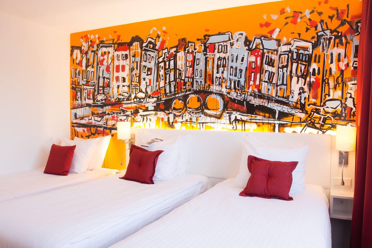 Habitación de hotel con dos camas, ropa de cama blanca, almohadas rojas y un cuadro abstracto grande en la pared.