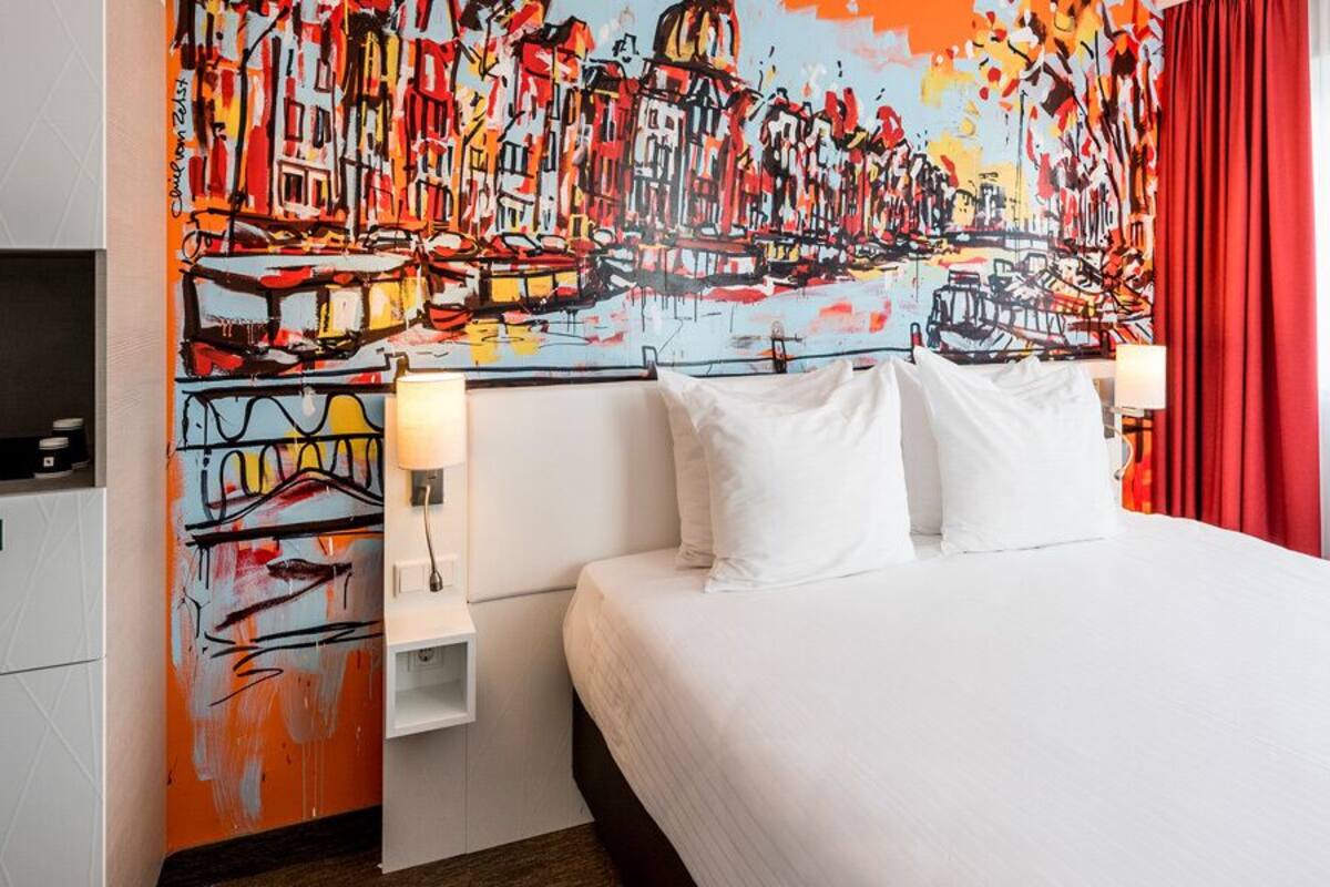 Una habitación de hotel con una cama grande, ropa de cama blanca, un cuadro abstracto colorido en la pared, una cortina roja y una mesita de noche pequeña con una lámpara.