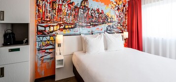 Une chambre d’h ôtel avec un grand lit, du linge de lit blanc, une peinture abstraite aux couleurs vives accrochée au mur, un rideau rouge et une petite table de chevet avec une lampe.