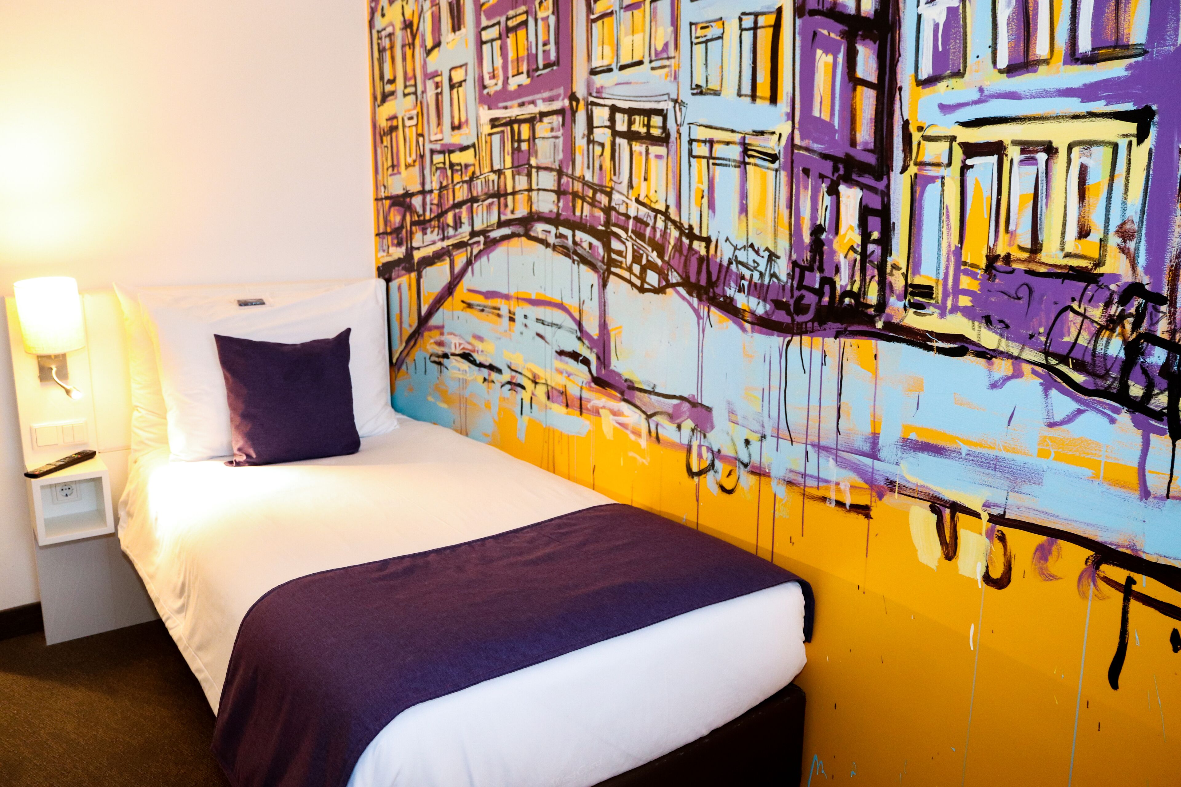 Habitació d'hotel amb llit, tauleta de nit, làmpada i un mural gran i colorit a la paret.