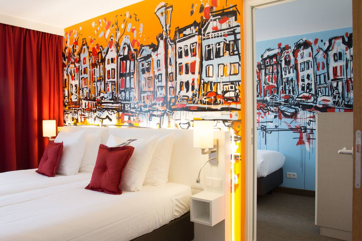 Una habitación de hotel con una cama grande, cortinas rojas, una mesita de noche blanca y un cuadro de un paisaje urbano en la pared.