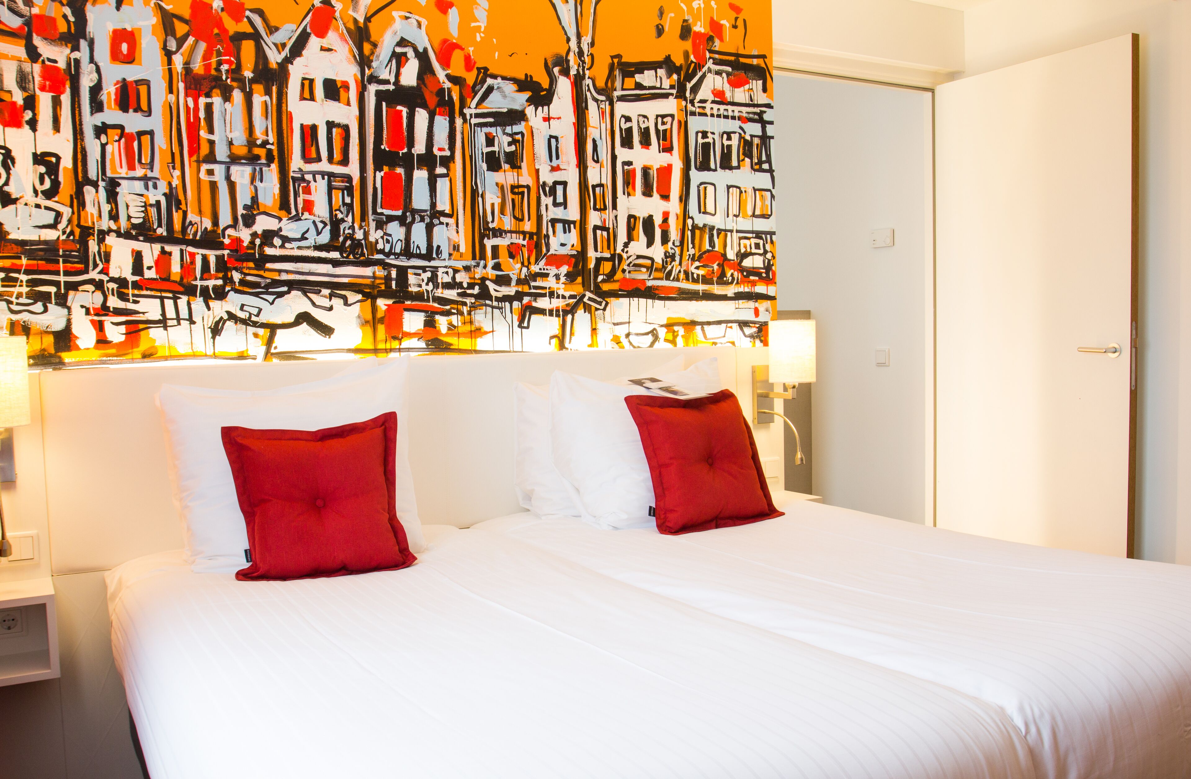 A hotel room with a large bed, two red pillows, and a vibrant abstract painting above the bed.