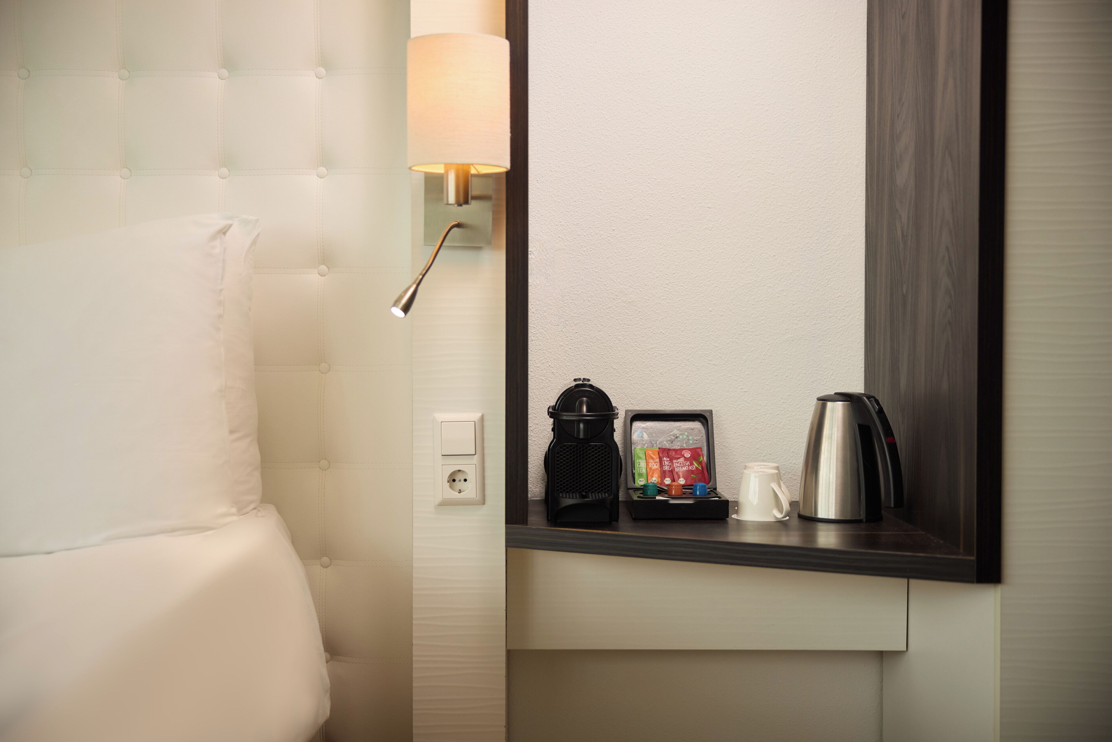 Un baño de hotel con un inodoro blanco, una toalla, una lámpara de pared, una cafetera y un juego de té.