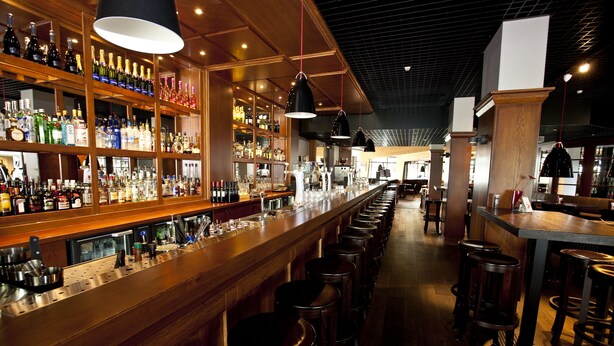 Een bar met houten planken vol met verschillende flessen, een lange toonbank met krukken, en hangende lampen.