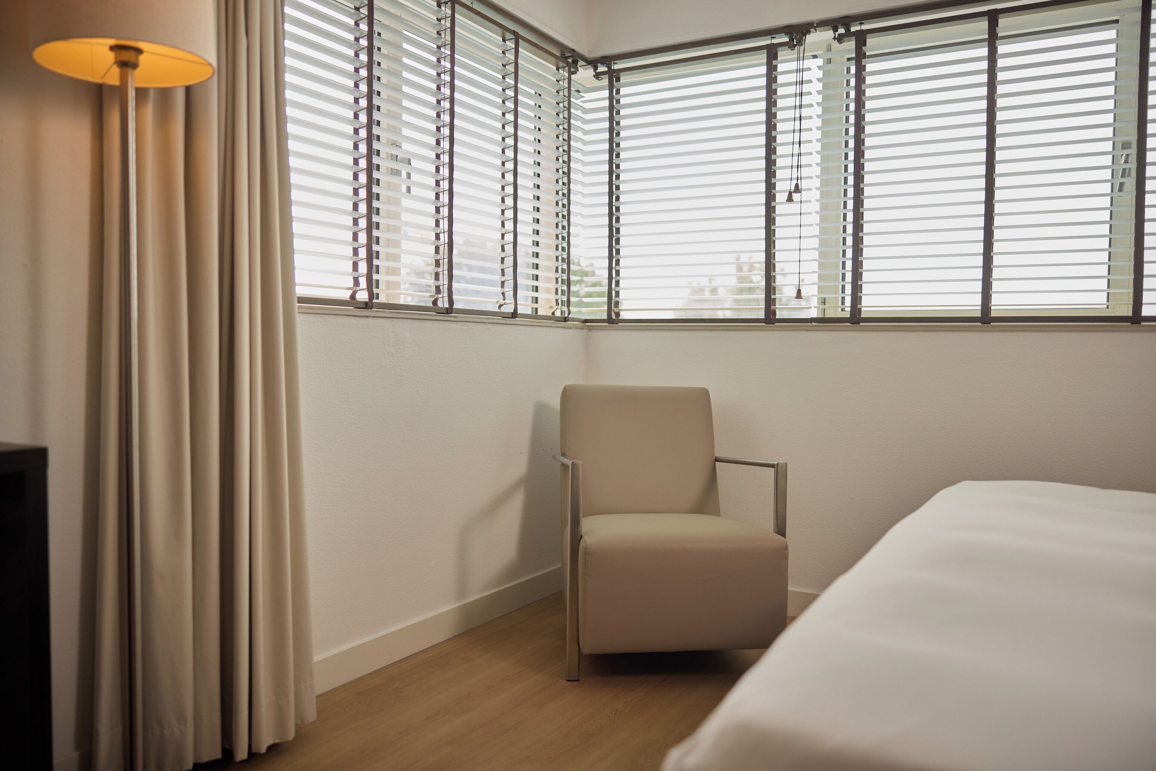 A hotel room with a bed, a chair, a lamp, and a window with blinds.