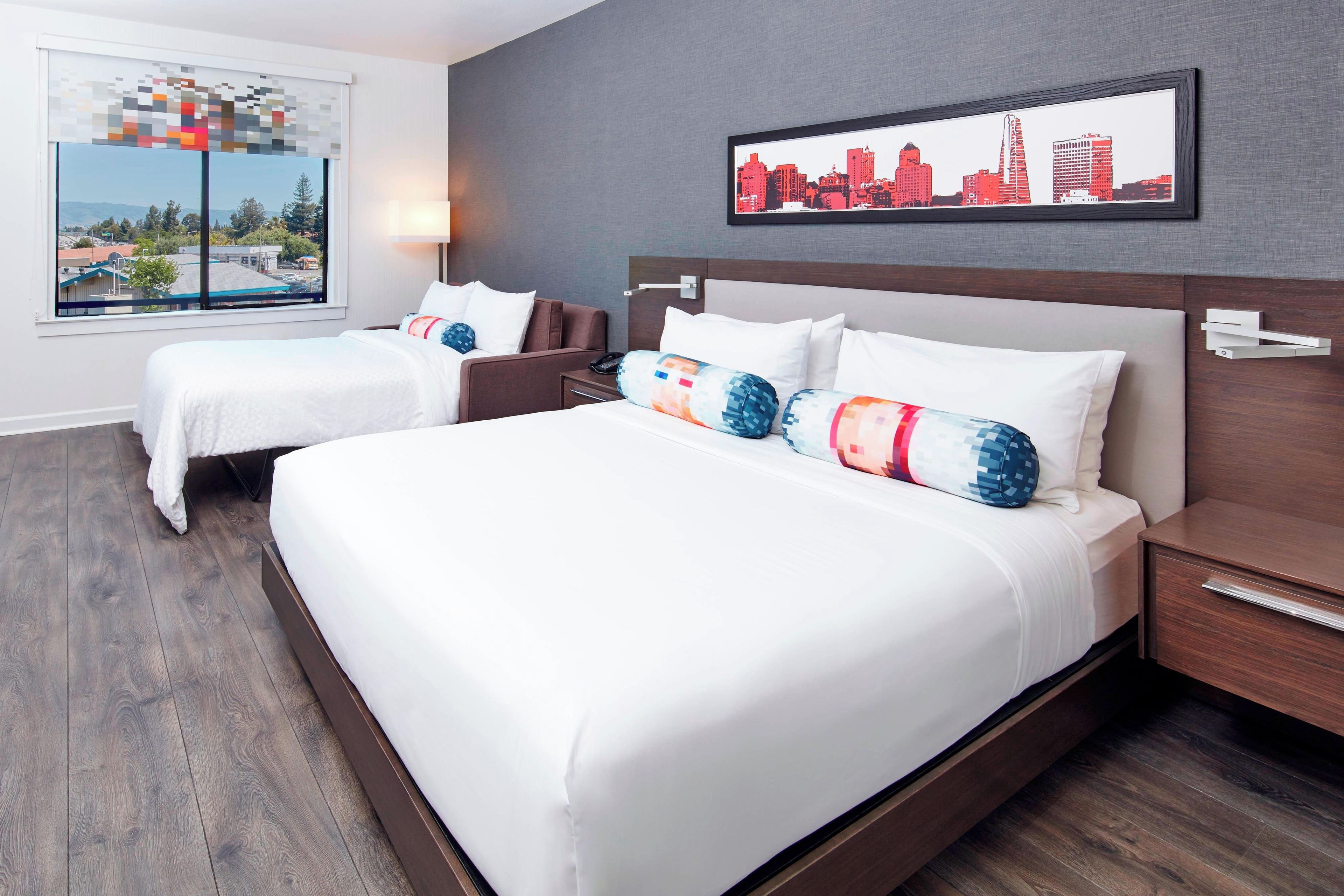 A hotel room with two beds, a cityscape artwork, and a window with a view.