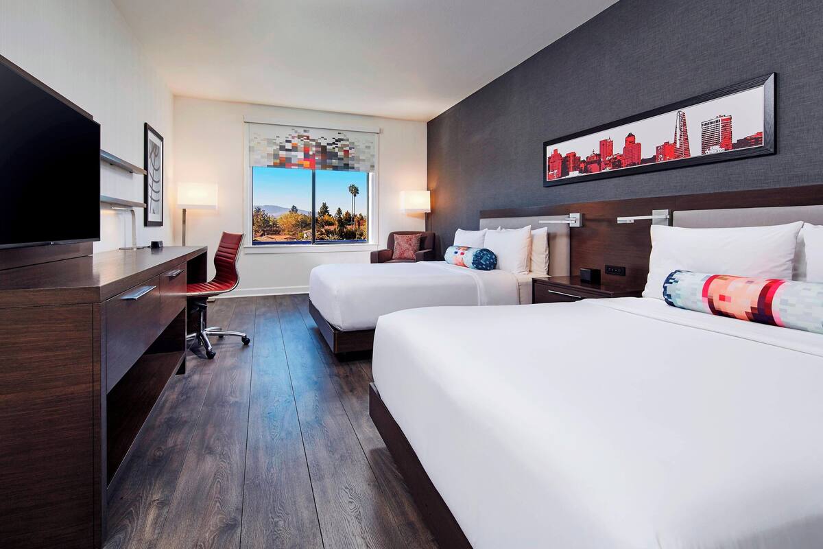 A hotel room with two beds, a desk, a TV, and a cityscape artwork on the wall.