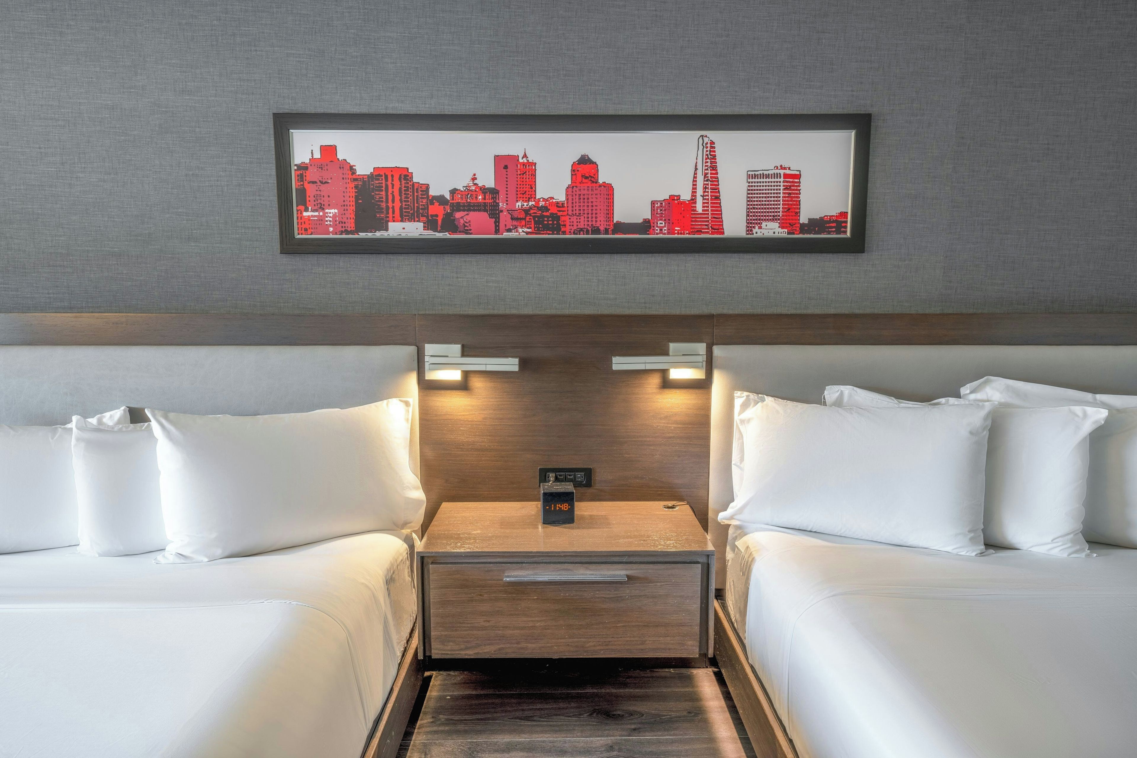 A hotel room with a cityscape artwork on the wall, a wooden headboard, two white beds, and a nightstand with a clock.