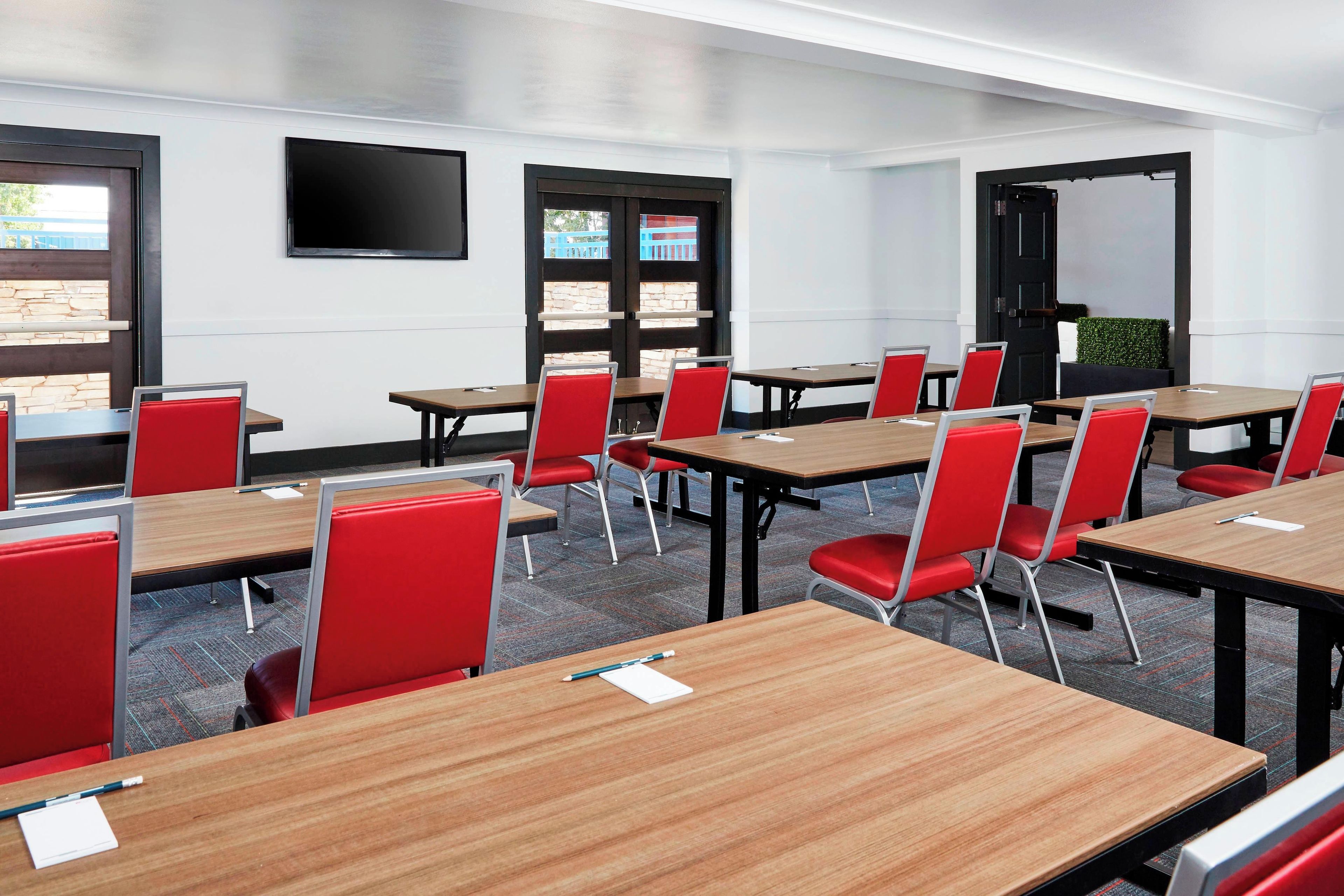 A conference room with tables and chairs, a flat-screen TV, windows, and a door.