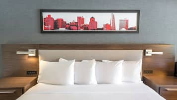 A neatly made bed with white linens and pillows, a framed cityscape artwork above it, and wooden headboard with bedside lamps.