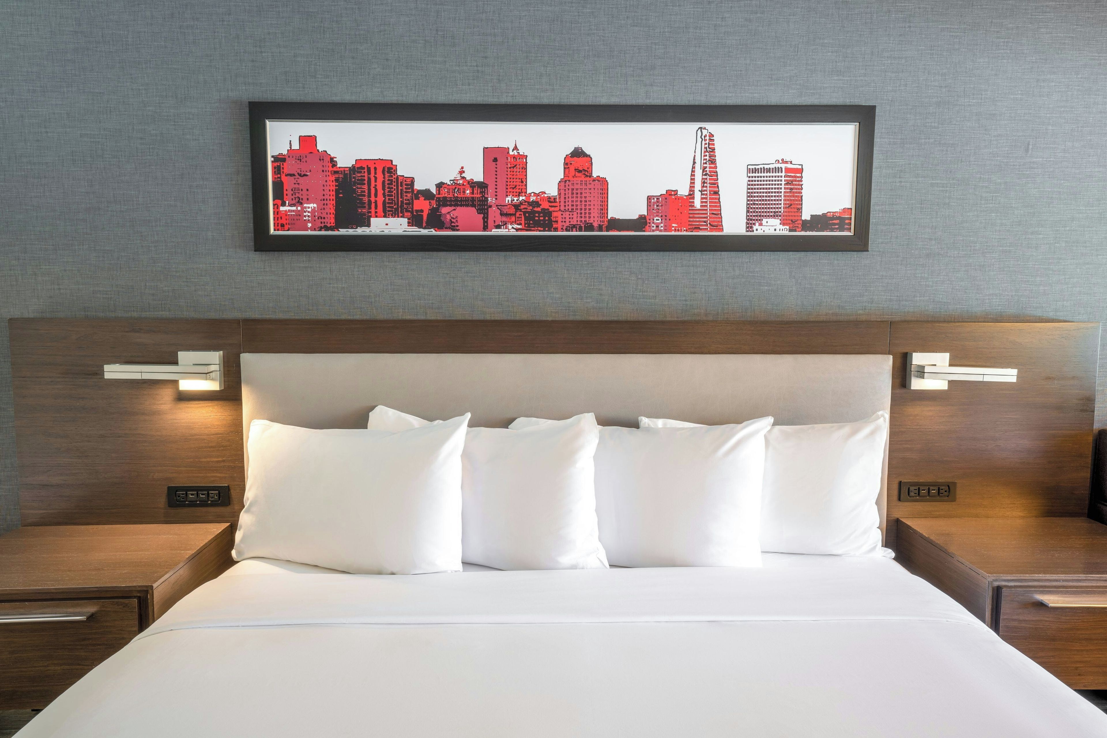 A hotel room with a large bed, white bedding, a framed cityscape artwork above the bed, and wooden headboard with bedside lamps.