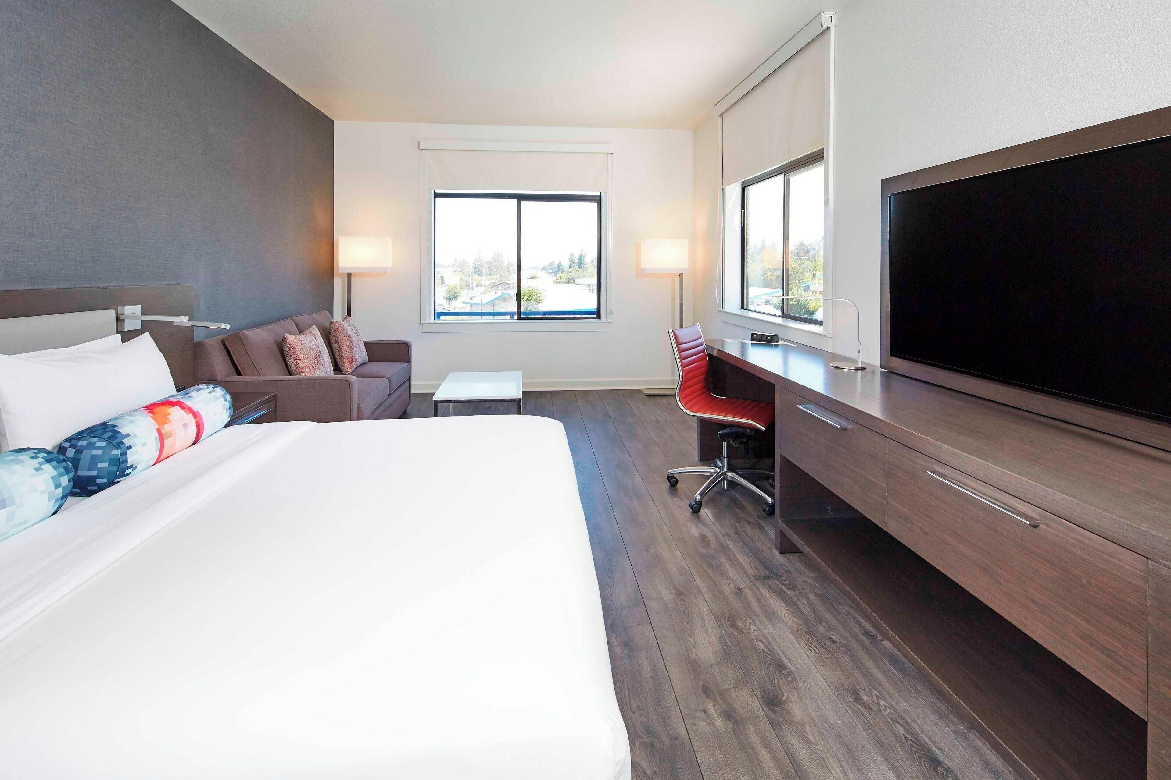 A hotel room with a large bed, a sofa, a desk with a TV, and a window with a view.