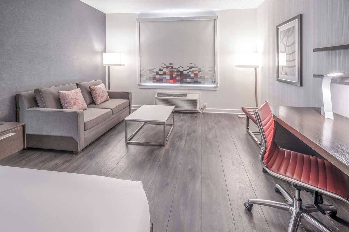 A modern, well-lit room with a grey sofa, a small table, a red office chair, and a large window with blinds.