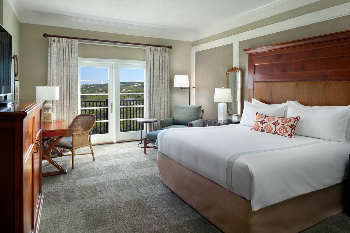 Ein Hotelzimmer mit einem großen Bett, einem Schreibtisch mit Stuhl, einem kleinen Tisch und Blick ins Freie durch eine Schiebetür.