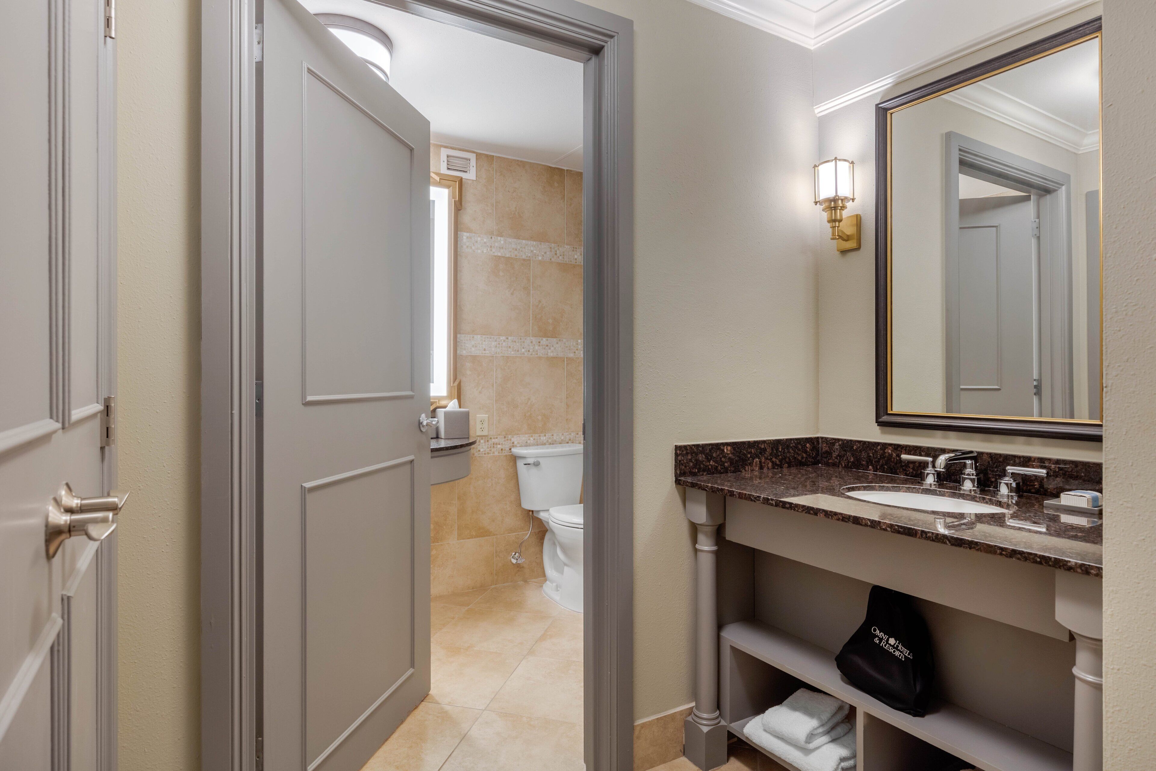Een moderne badkamer met een marmeren wastafel, een grote spiegel en een toilet dat door een open deur zichtbaar is.