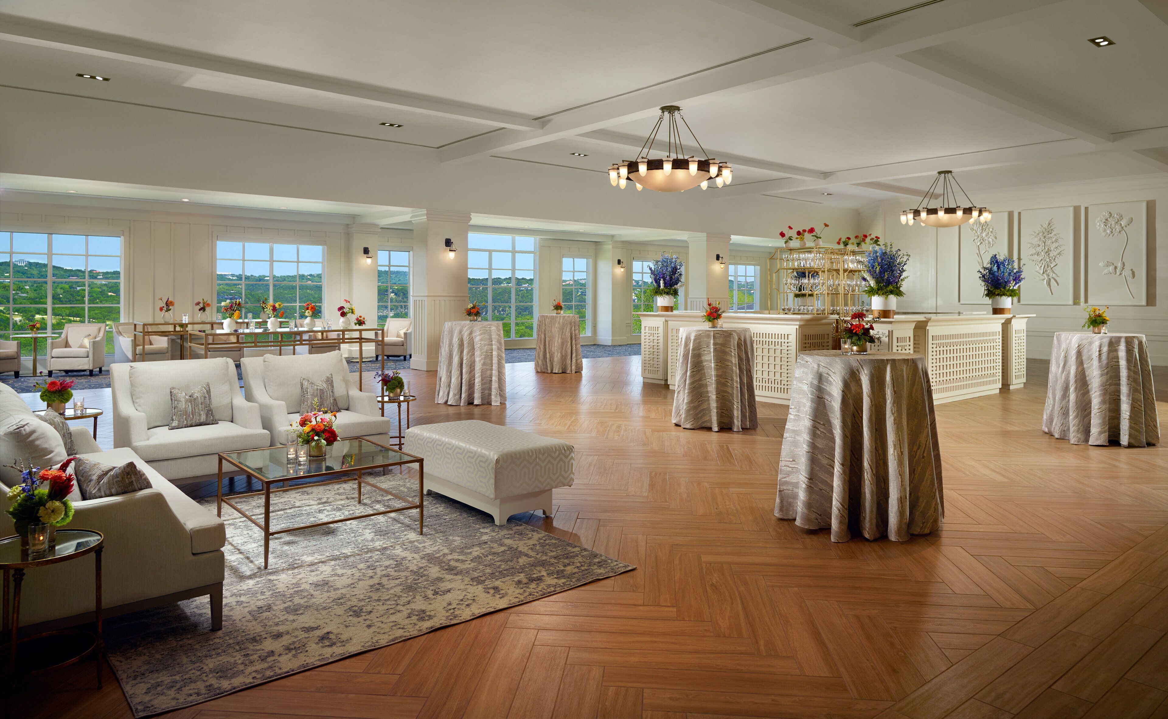 A spacious hotel lobby with a high ceiling, large windows offering a view of the outdoors, and a central area with tables and chairs.