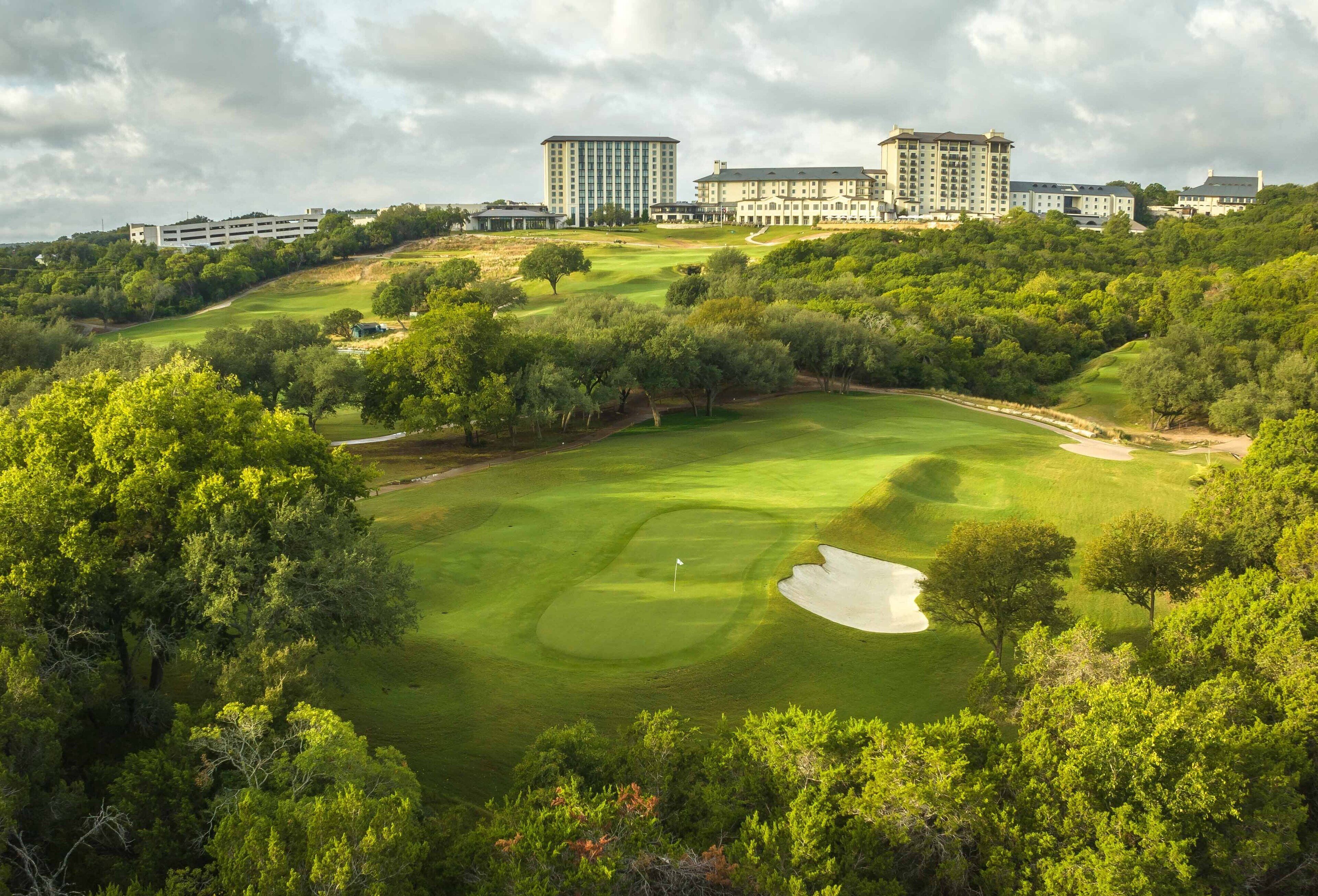 A golf course with a sand bunker and a flagstick, surrounded by dense trees and multiple buildings in the background.