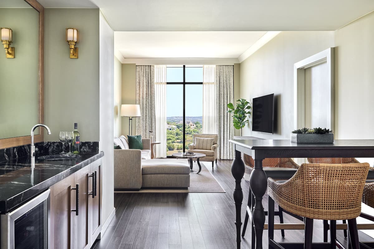 Ein modernes Hotelzimmer mit Küchenbereich, einem Esstisch mit Stühlen, einer Couch und einem großen Fenster mit Blick auf die Stadt.