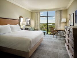A hotel room with a large bed, a desk, a chair, and a dresser. There is a window with a view of trees and buildings.