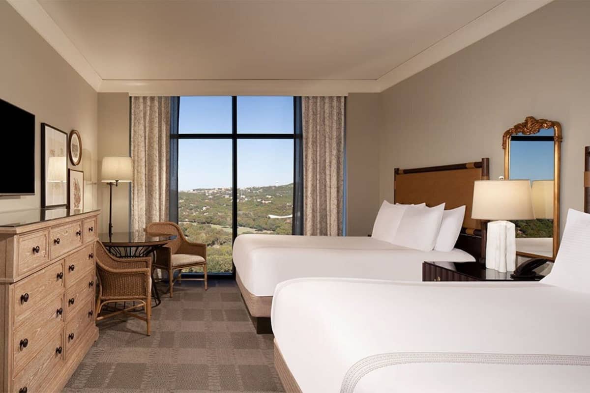 Ein Hotelzimmer mit zwei Betten, einem Schreibtisch, einem Stuhl, einem Fernseher und einem großen Fenster mit Blick auf die Landschaft.