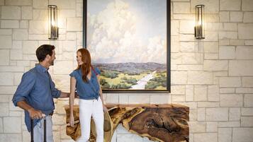 A couple standing in front of a painting with a landscape view, a wooden table, and stone walls.