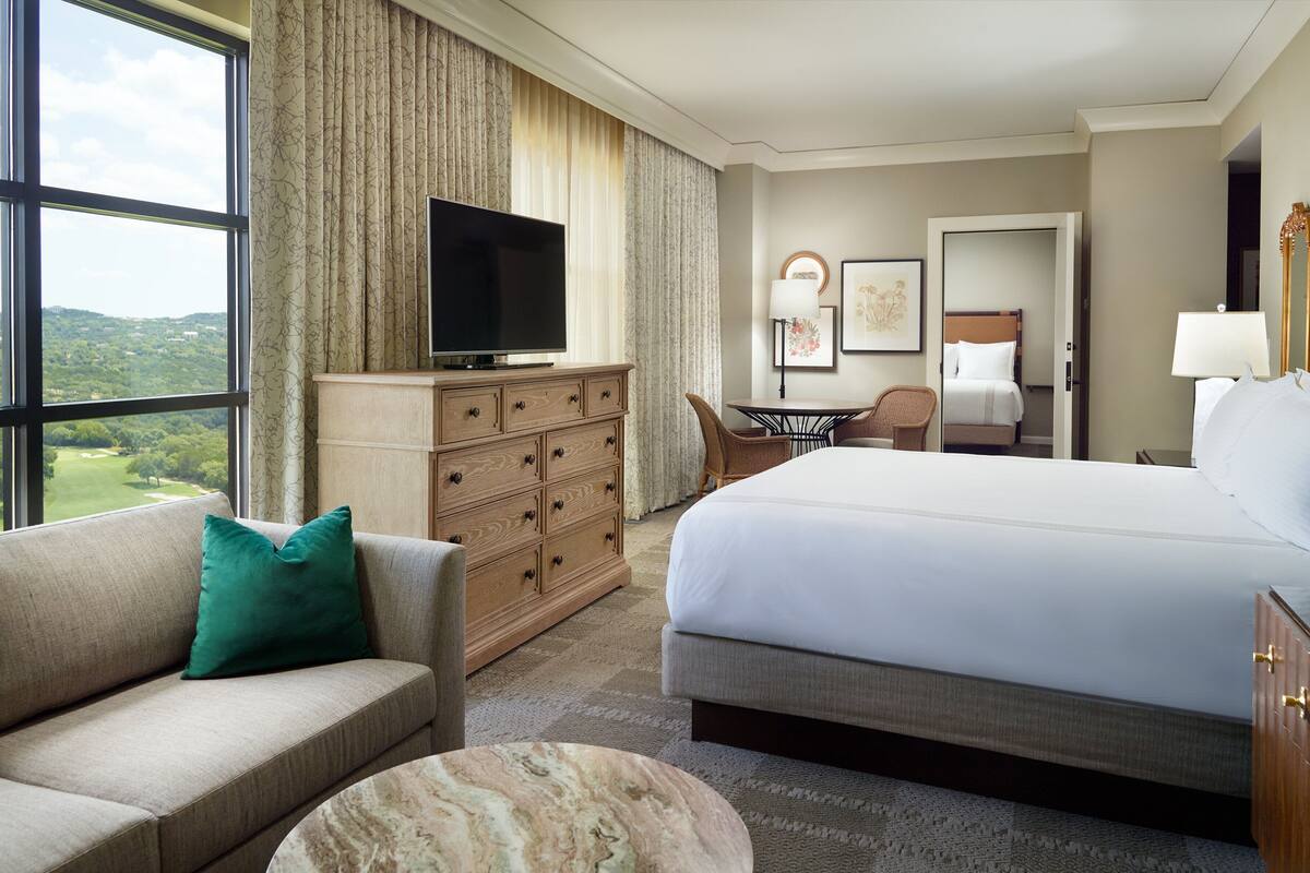 Ein Hotelzimmer mit einem großen Bett, einem Sofa, einem Fernseher, einem Schminktisch und Blick ins Freie.