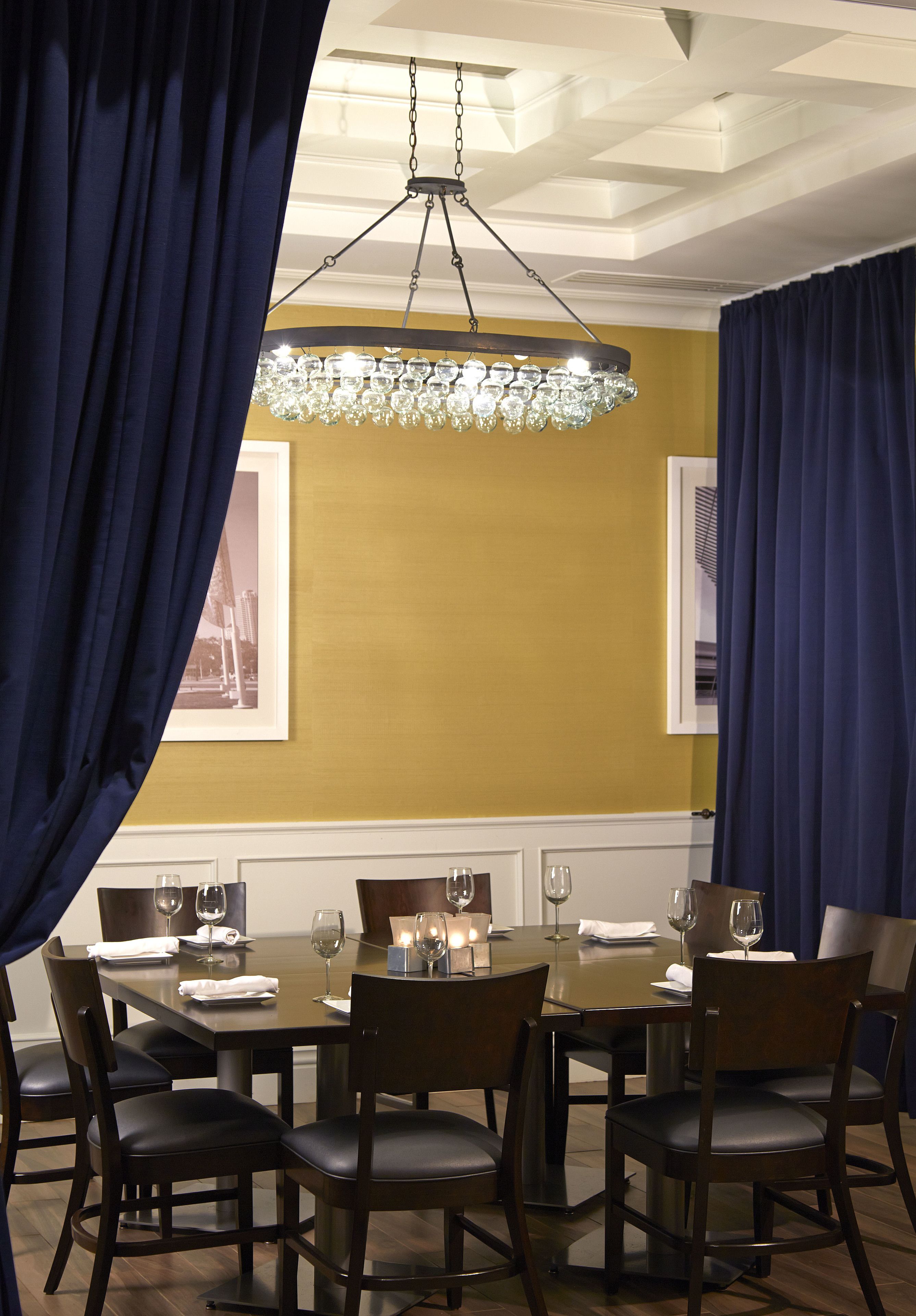 Comedor con una mesa dispuesta para cuatro personas, rodeada de sillas, con una araña colgante y cortinas a ambos lados.