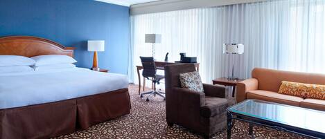 A hotel room with a large bed, a sofa, a glass coffee table, a desk with a lamp, and a chair.