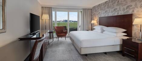 A hotel room with a large bed, a desk with a TV, a chair, and a view of a green field through the window.
