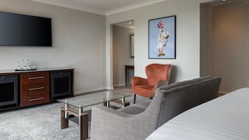 A hotel room with a bed, a grey armchair, a red armchair, a glass coffee table, a wooden dresser, a flat-screen TV, and a framed artwork on the wall.