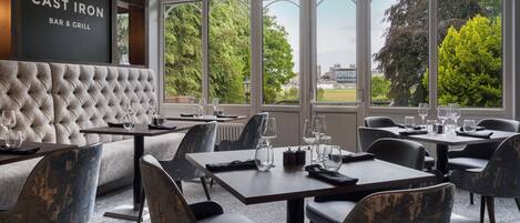 A restaurant with a bar and grill, featuring a tufted headboard, large windows with a view of trees and buildings, and a mix of modern and classic furniture.