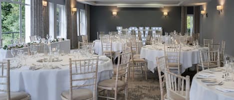 Un salón de banquetes con mesas redondas dispuestas para comer, ventanas amplias e iluminación empotrada.