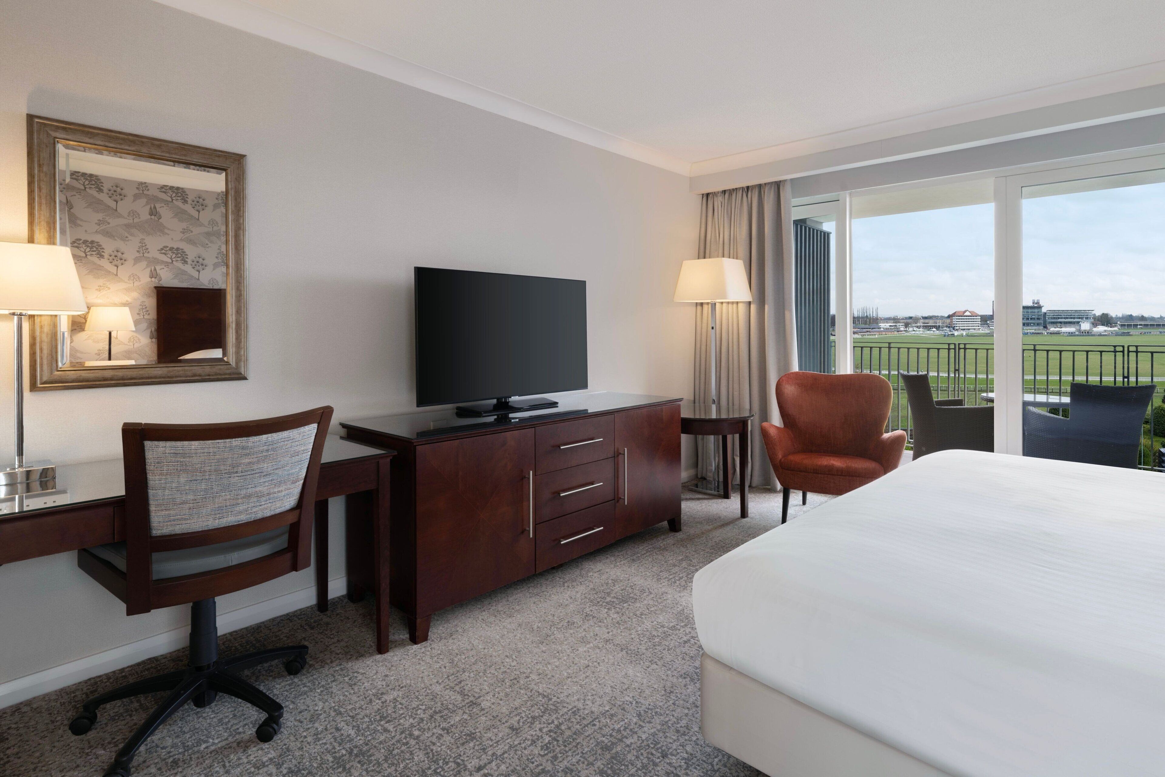A hotel room with a bed, a desk with a chair, a television, and a view of a sports field through the window.