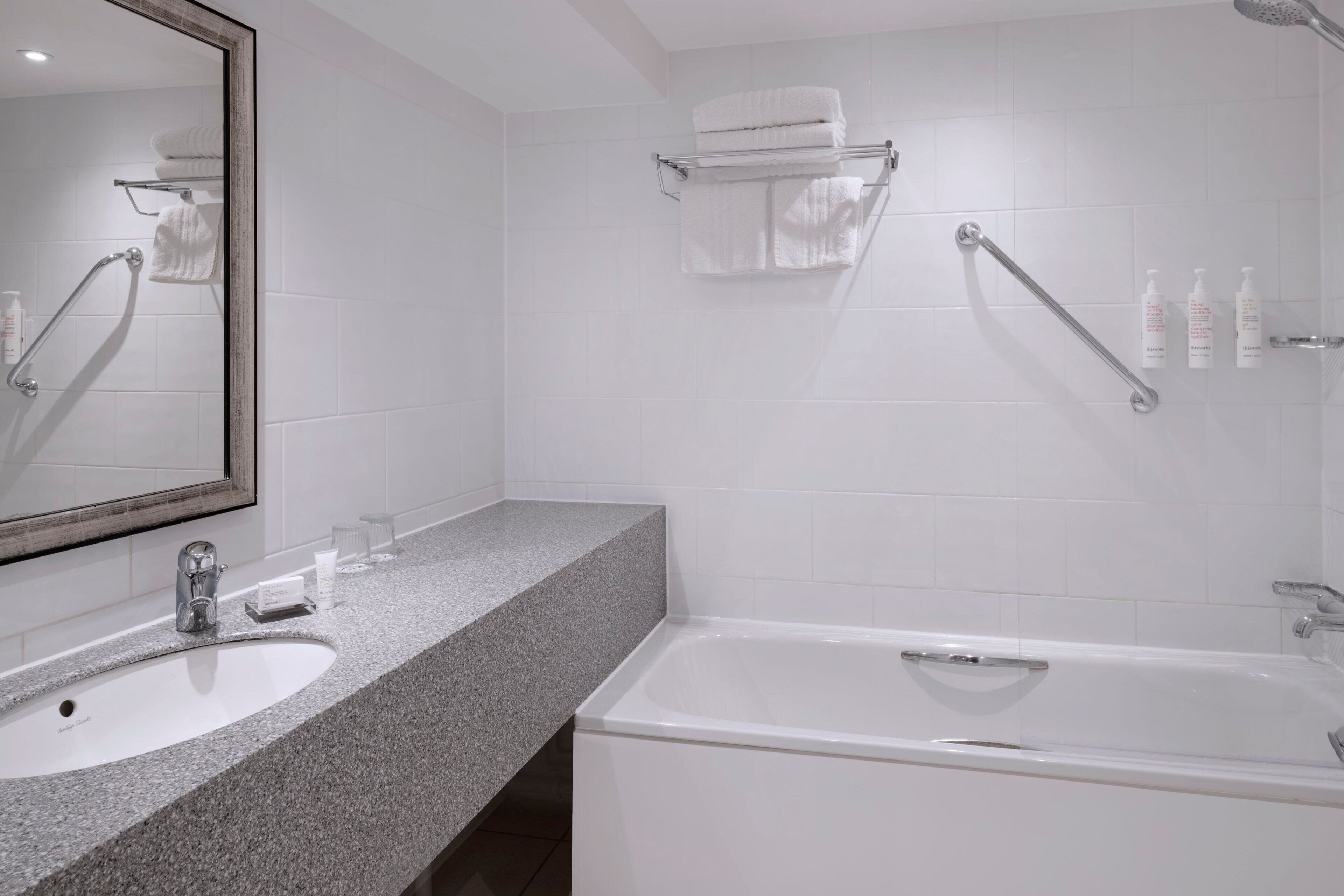 Une salle de bain moderne avec un lavabo surmonté d’un plan de travail en granit, un grand miroir, une baignoire blanche et un porte-serviettes.