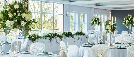 A banquet hall with round tables set for a formal event, adorned with floral centerpieces and white tablecloths.