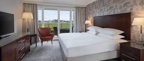 A hotel room with a large bed, a TV, a chair, and a balcony with a view of a field.